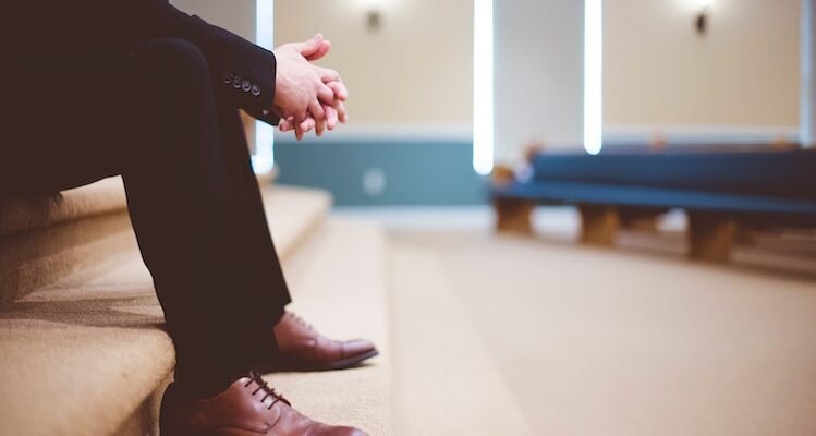 man in black pants and pair of brown leather lace-up shoes sitting on brown carpeted stairs inside room